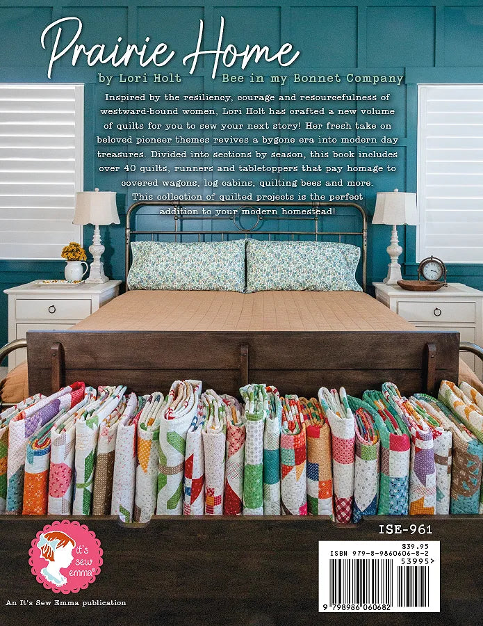 A neatly made bed with blue-green walls and two side tables beneath windows. At the foot, a bench is stacked with colorful quilts inspired by Checker Distributor’s Prairie Home Quilt Book. Text above reads “Prairie Home.”.