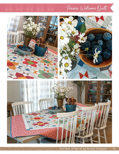 A bright, cozy room features a dining table with a colorful quilt from the Prairie Home Quilt Book by Checker Distributor, white wooden chairs, a metal jug of white flowers, and bowls of blueberries.