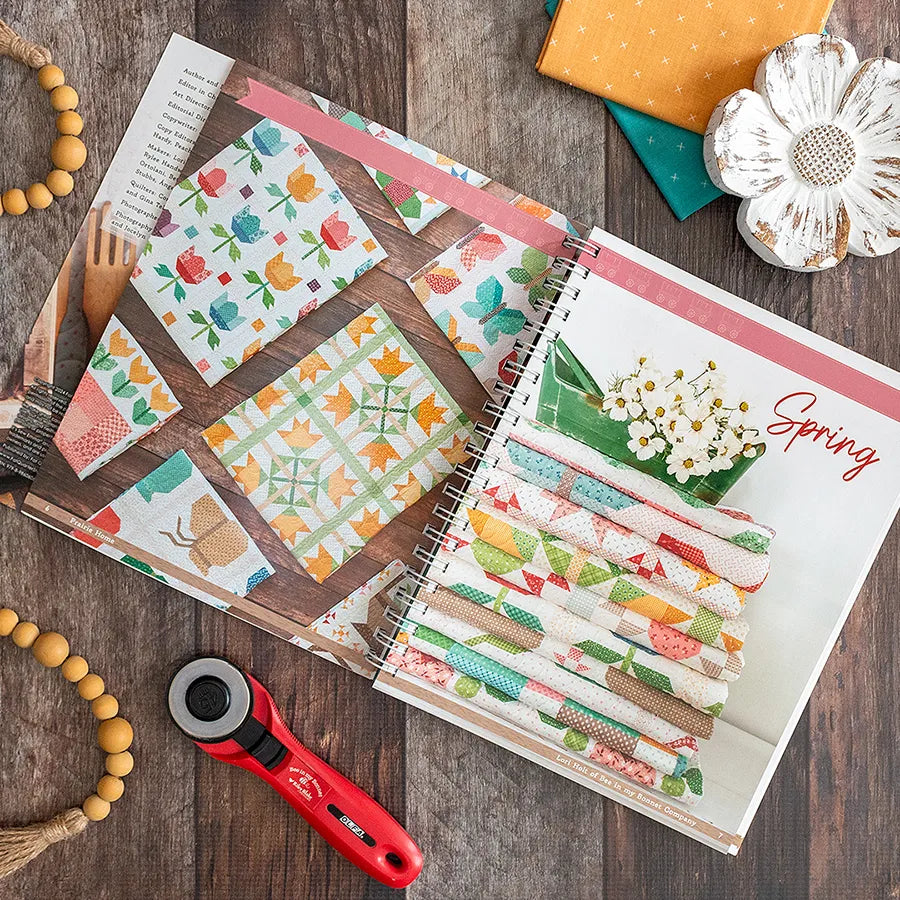 A Prairie Home Quilt Book by Checker Distributor is open to bright quilting patterns with folded fabric, a rotary cutter, beaded bracelet, yellow and teal squares, and a white flower, all arranged on a wooden surface.