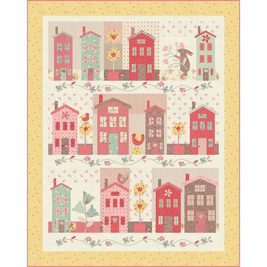 The Sunshine Lane Boxed Quilt Kit by United Notions features charming house rows in pink, red, teal, and beige, with floral, bird, rooster, and patchwork motifs framed by a yellow floral border.