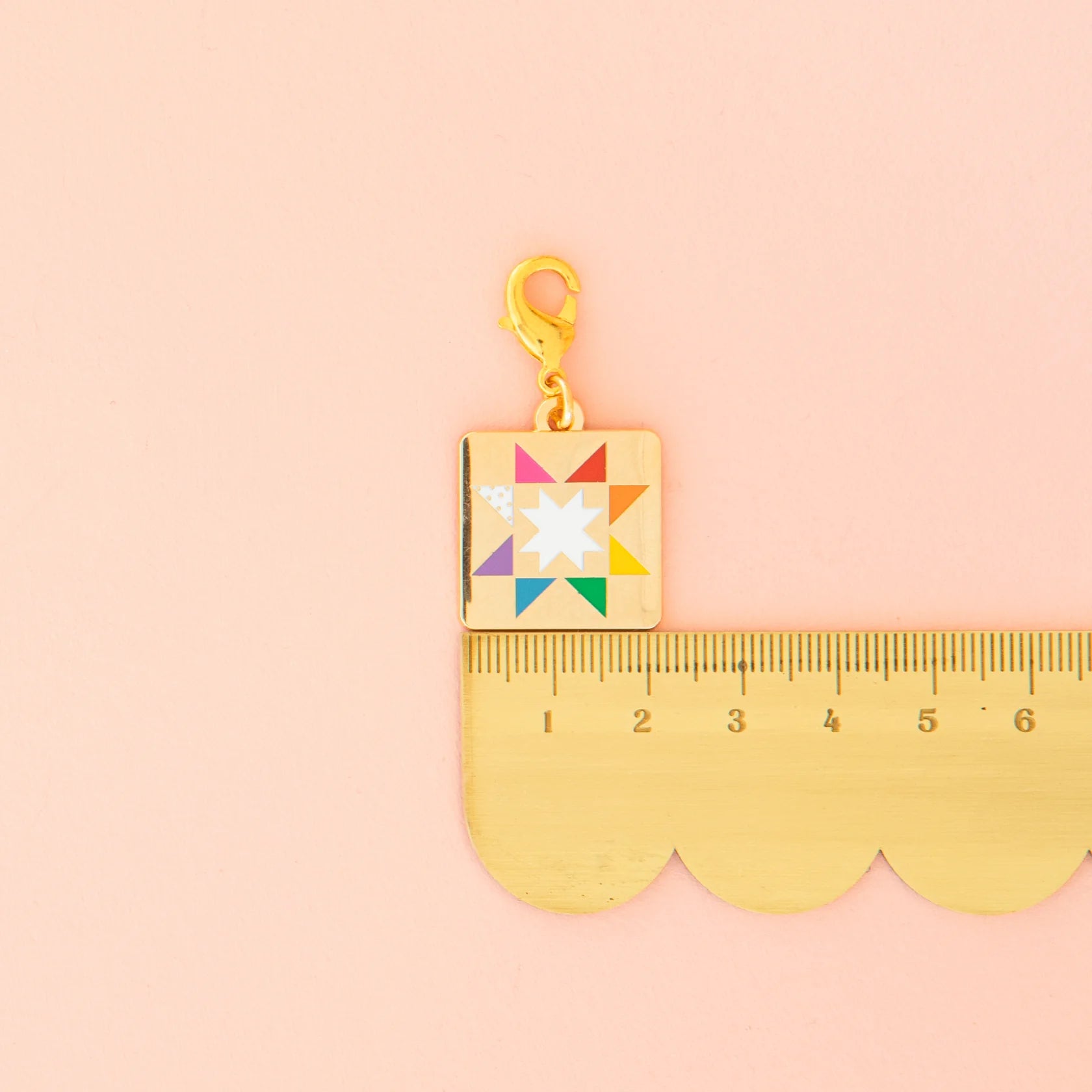 The Sarah Hearts Quilt Star Zipper Charm, featuring a colorful quilt star design and gold clasp, is shown beside a gold ruler on a pastel pink background. The zipper charm measures about 2 centimeters long.