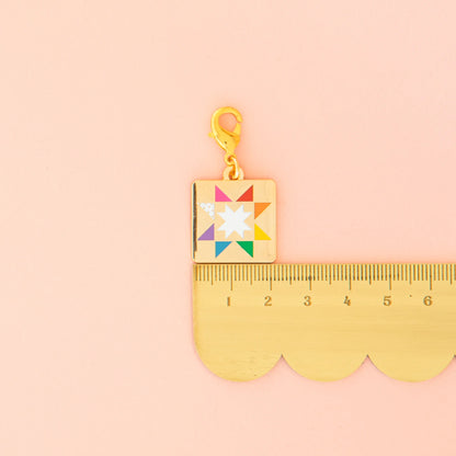 The Sarah Hearts Quilt Star Zipper Charm, featuring a colorful quilt star design and gold clasp, is shown beside a gold ruler on a pastel pink background. The zipper charm measures about 2 centimeters long.