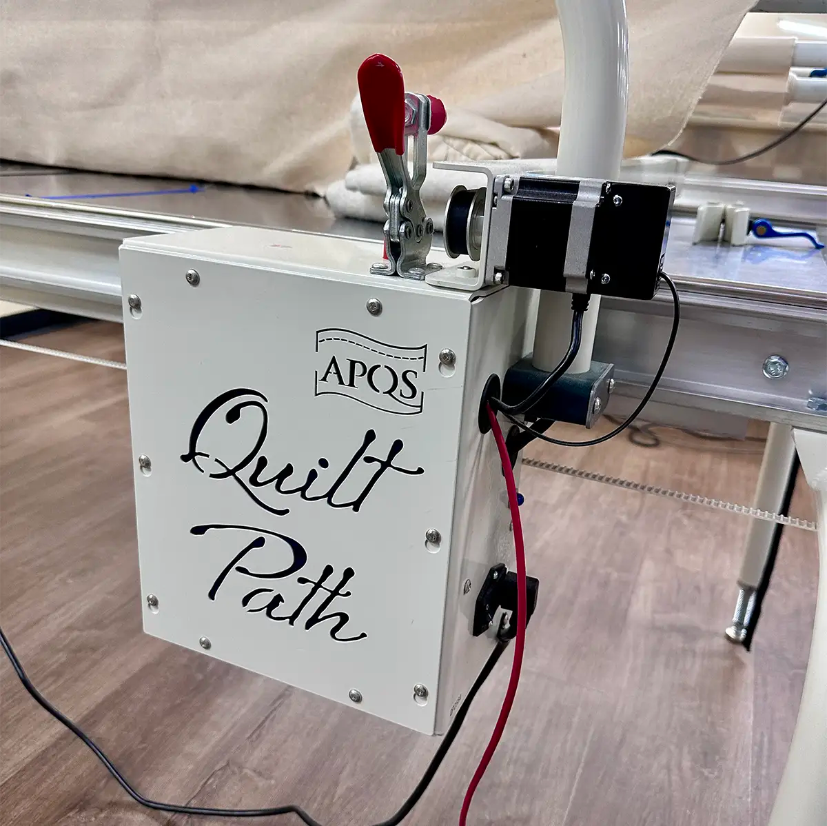 A close-up of the APQS Quilt Path Essentials Robot Computer attached to a quilting frame, showing its labeled white control box, red lever, motor, and wires with a wooden floor in the background.