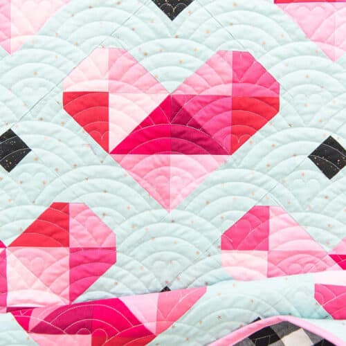 A quilt featuring a geometric heart pattern made from pink and red triangles on a pale blue background with an arched, scallop-like quilted stitching design.