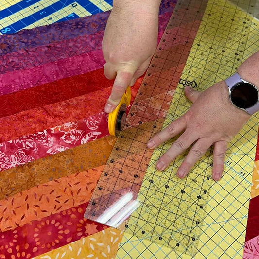 A person uses a rotary cutter and clear ruler to cut strips of colorful fabric on a gridded mat, possibly preparing Scrappy Binding for quilt finishing. A smartwatch is visible on their left wrist.