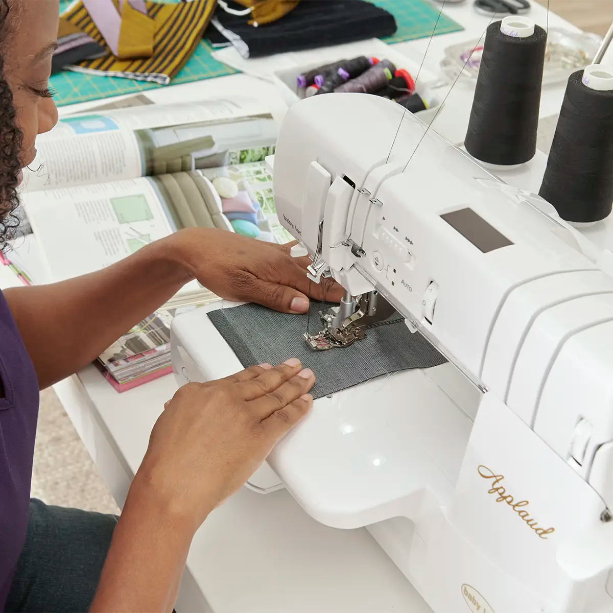 A person sews denim on a Quilted Joy Baby Lock Serger Pixie Playdate with black thread, surrounded by sewing supplies and an open book—great inspiration for anyone taking a serger class or learning to thread a serger.