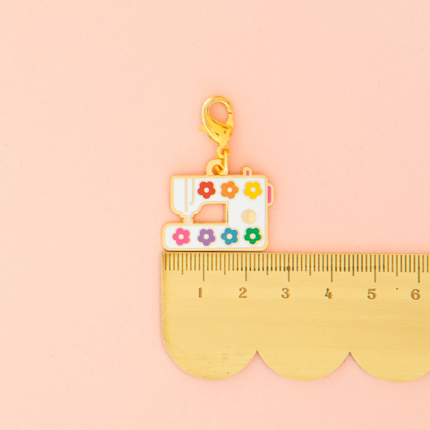 The Sarah Hearts Sewing Machine Zipper Charm features colorful flower details and hangs from a gold clasp. Shown beside a golden ruler, this accessory charm rests on a light pink background.