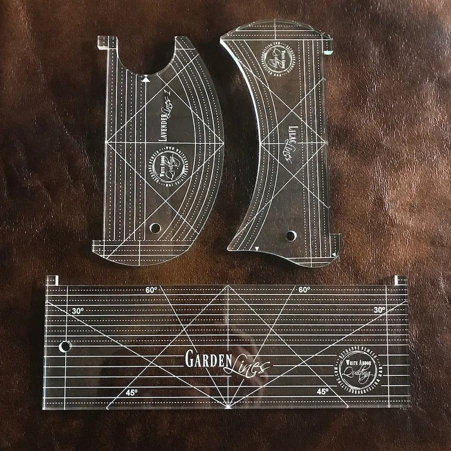 The Stitch in the Ditch Collection Ruler Set by White Arbor Quilting features three transparent rulers with white geometric markings and “Garden Lines” text, including curved options for continuous curve quilting, shown on a brown leather surface.