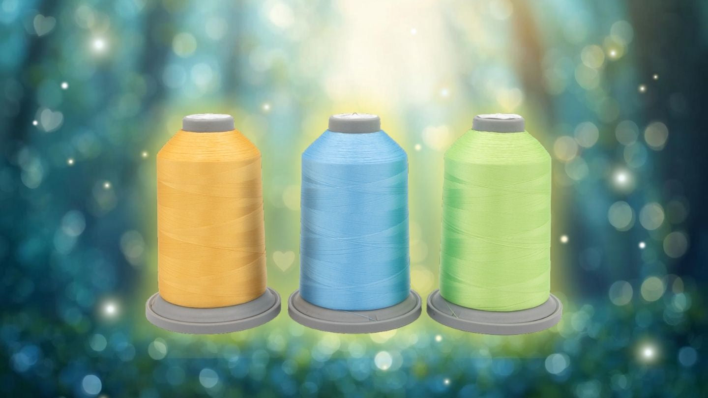 Three large spools from the Sugar Stitches Glide Thread of the Month Bundle by Hab+Dash by Fil Tec—yellow, blue, and green—are displayed side by side against a softly blurred background with sunlight and greenery.