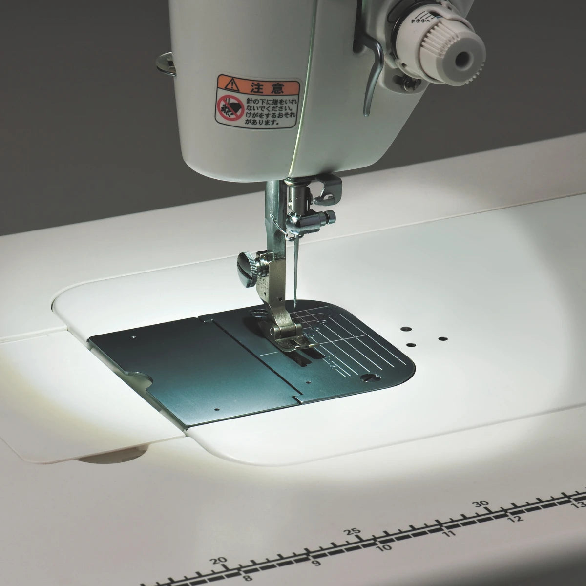 The needle area of the JUKI TL-15 lit by its LED against a dark background, with the inch markings along the front edge of the bed catching the light.
