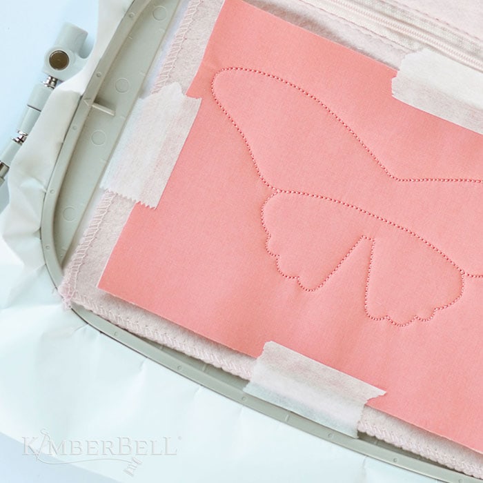 image of embroidery hope with a butterfly sewn into pink fabric and Kimberbell Paper Tape holding the fabric in place