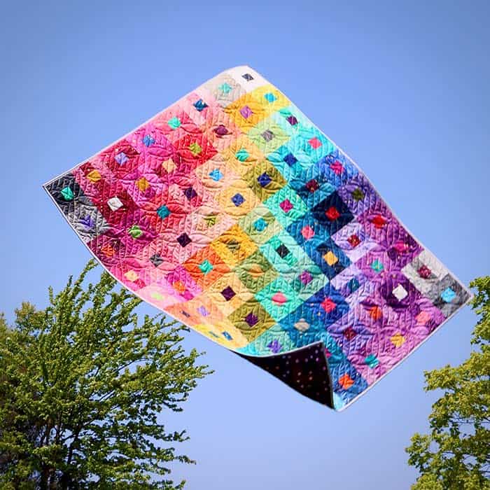 Image of Ombre Galaxy Metallic Jelly Roll quilt tossed into the air with a blue sky and trees in the background